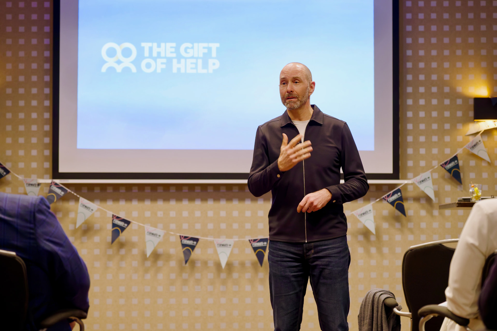 Ashley Usiskin speaking with his hand on his chest, The Gift of Help on the screen behind him