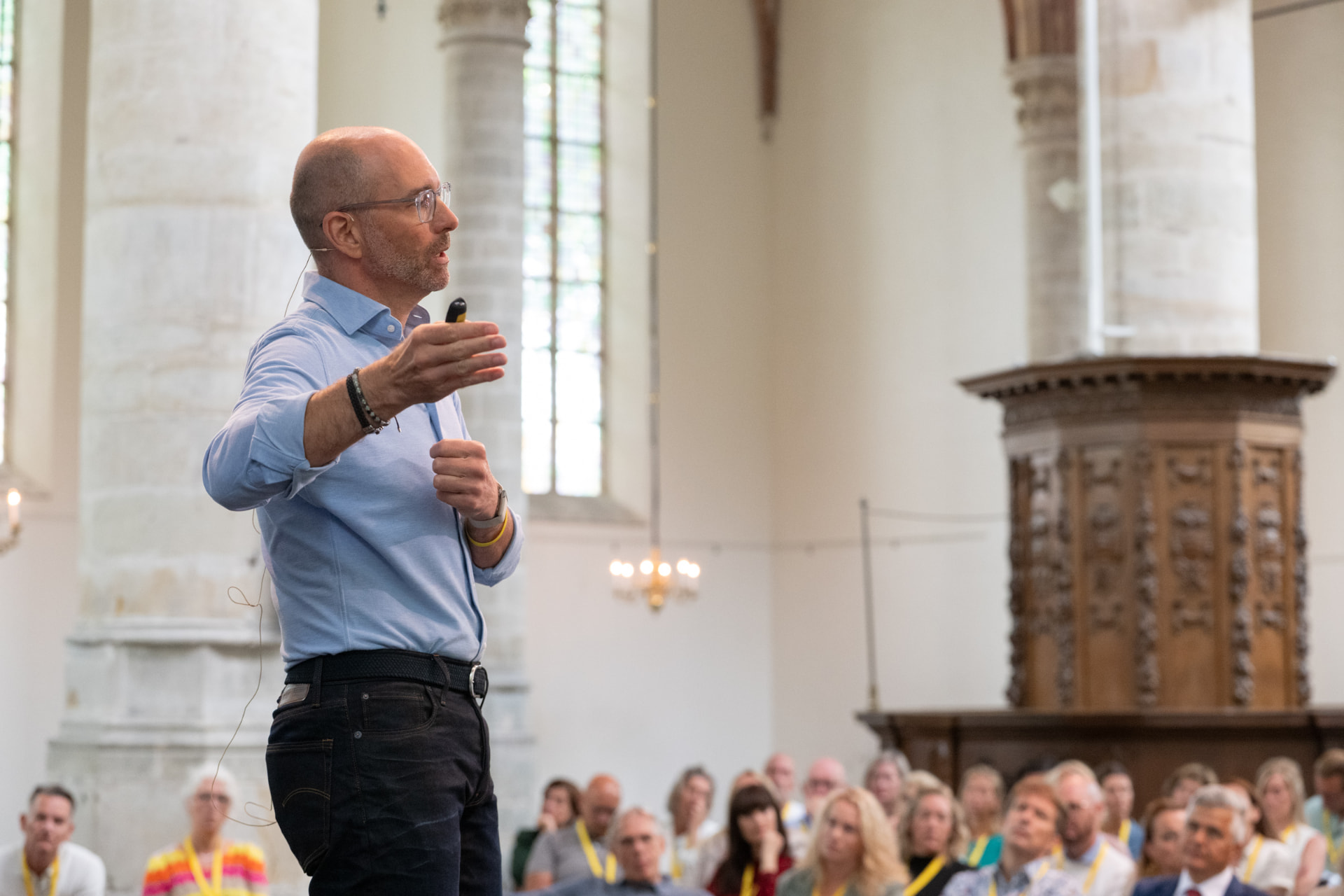 Ashley Usiskin speaking in profile with an outstretched hand and slide controller, addressing a seated audience in a large venue